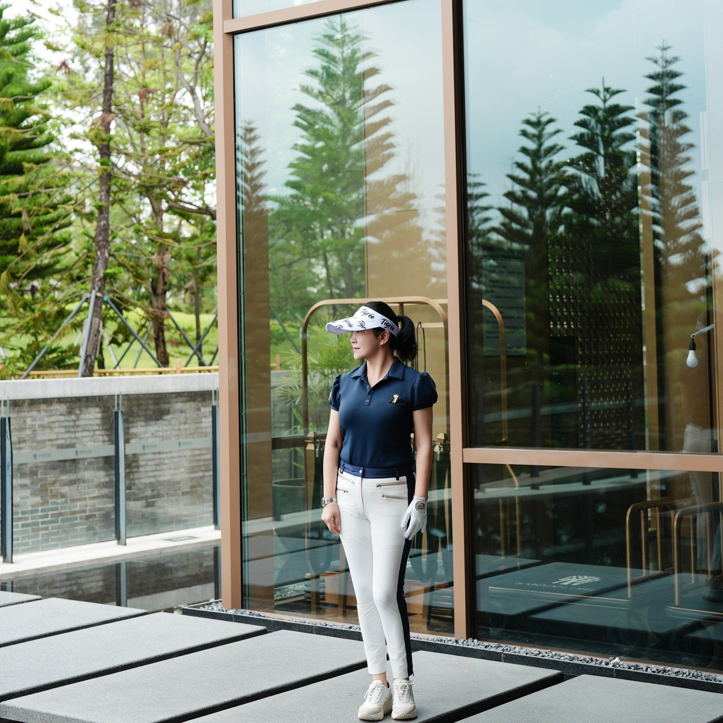 A woman wearing a navy blue polo shirt, white pants, and a white visor stands on a modern outdoor walkway in front of a glass-walled building with trees reflected in the windows.