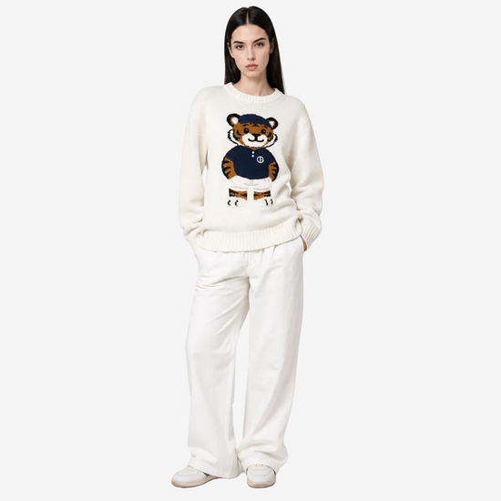 A model wearing a cream-colored knit sweater with a cartoon tiger graphic on the chest, paired with matching cream wide-leg trousers and white sneakers, set against a plain light gray studio background.