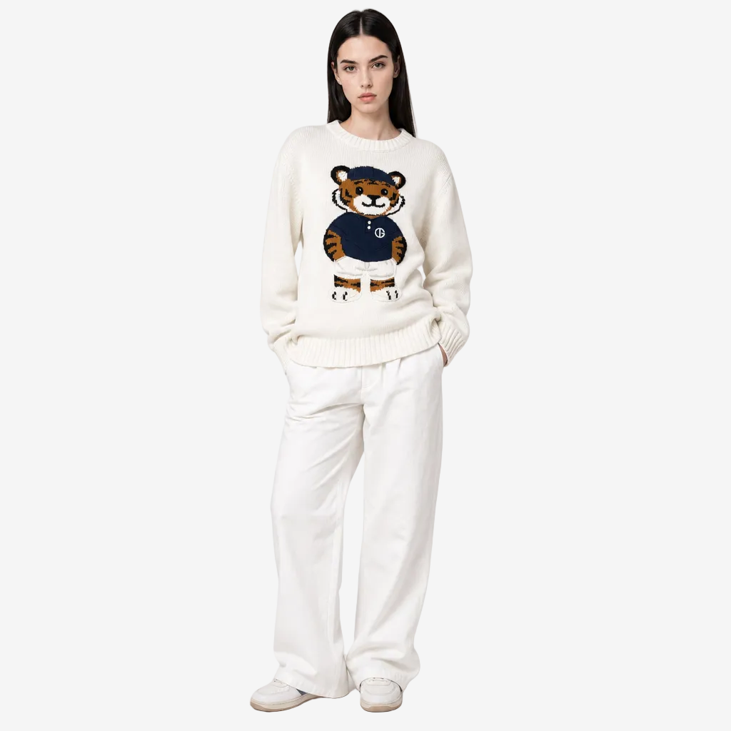 A model wearing a cream-colored knit sweater with a cartoon tiger graphic on the chest, paired with matching cream wide-leg trousers and white sneakers, set against a plain light gray studio background.
