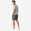 A man wearing a light sage green t-shirt, gray and black camouflage shorts, and white sneakers, standing in profile against a light gray studio background.