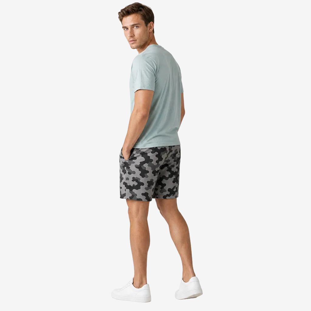 A man wearing a light sage green t-shirt, gray and black camouflage shorts, and white sneakers, standing in profile against a light gray studio background.
