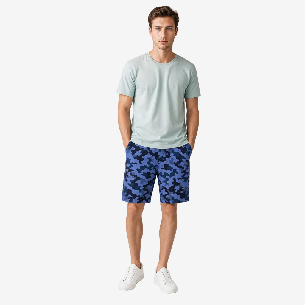A man modeling a light sage green crewneck t-shirt and blue camouflage shorts against a plain light gray studio background.