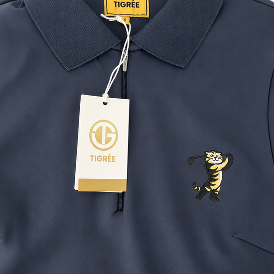 A close-up of a navy blue polo shirt with a yellow TIGRÉE neck label, a white hangtag, and a yellow embroidered tiger mascot on the chest.