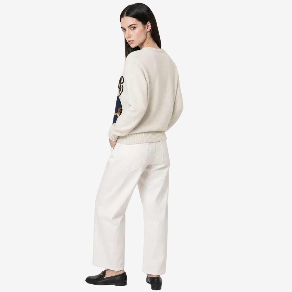 Woman wearing a cream sweater and white pants on a plain background