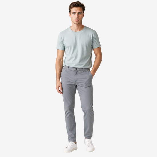 A man modeling a light sage green crewneck t-shirt and gray slim-fit trousers with white sneakers, standing against a plain white studio background.