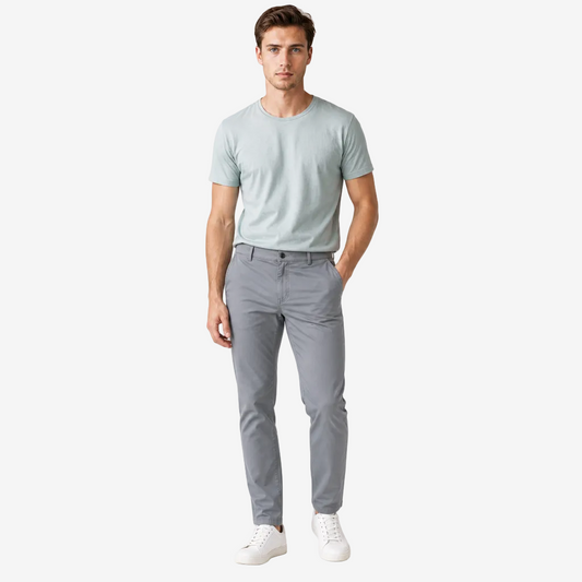 A man modeling a light sage green crewneck t-shirt and gray slim-fit trousers with white sneakers, standing against a plain white studio background.