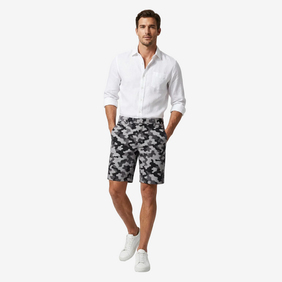 A man modeling a white long-sleeve button-down shirt and black-and-white camouflage shorts, paired with white sneakers, against a light gray studio background.