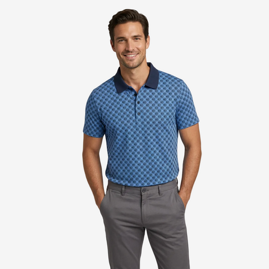 A man wearing a blue short-sleeve polo shirt with a geometric pattern and a dark navy collar, paired with gray trousers, standing against a light gray studio background.