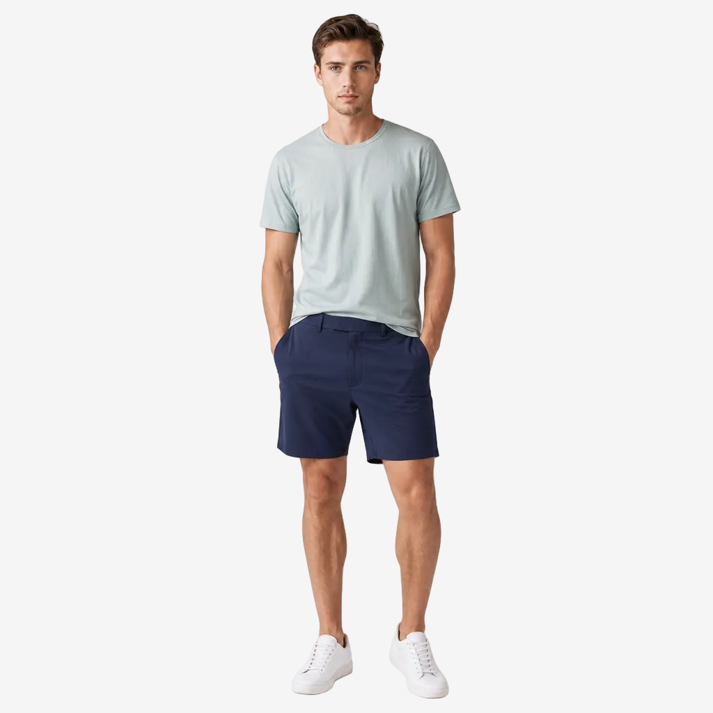 A man modeling a light sage green crewneck t-shirt and navy blue shorts, paired with white sneakers, standing against a neutral light gray studio background.