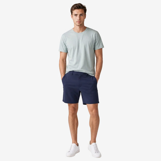 A man modeling a light sage green crewneck t-shirt and navy blue shorts, paired with white sneakers, standing against a neutral light gray studio background.