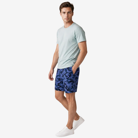 A man modeling a light sage green crewneck t-shirt and blue camouflage shorts, paired with white sneakers, against a plain light gray studio background.