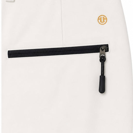 Close-up of off-white trousers showing a front zip pocket with a black zipper and a small gold circular logo patch near the waist.