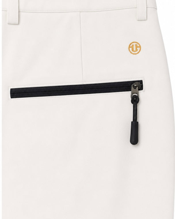 Close-up of off-white trousers showing a front zip pocket with a black zipper and a small gold circular logo patch near the waist.