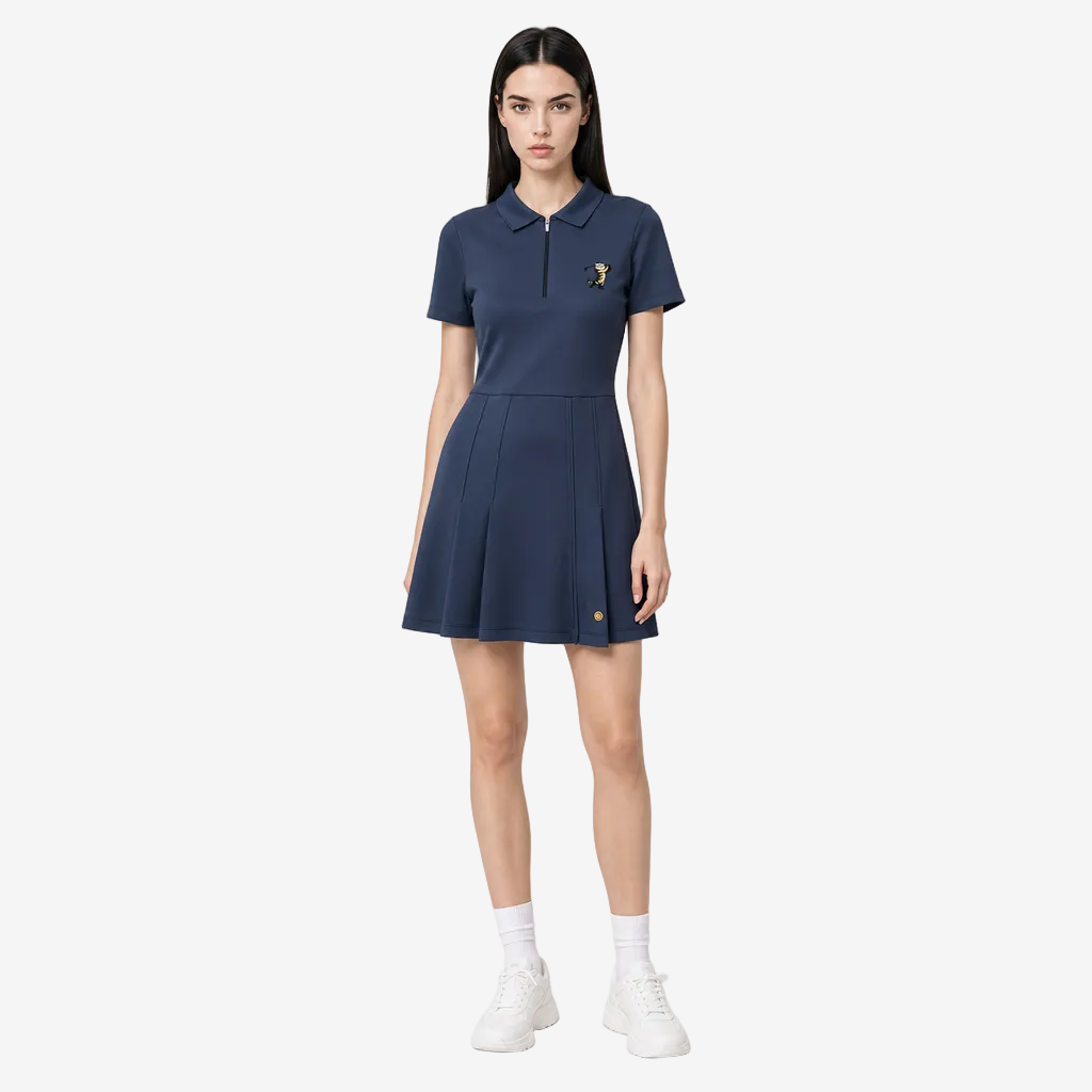 A woman modeling a navy blue short-sleeve polo dress with a small gold embroidered logo on the chest, paired with white socks and white sneakers, against a light gray studio background.