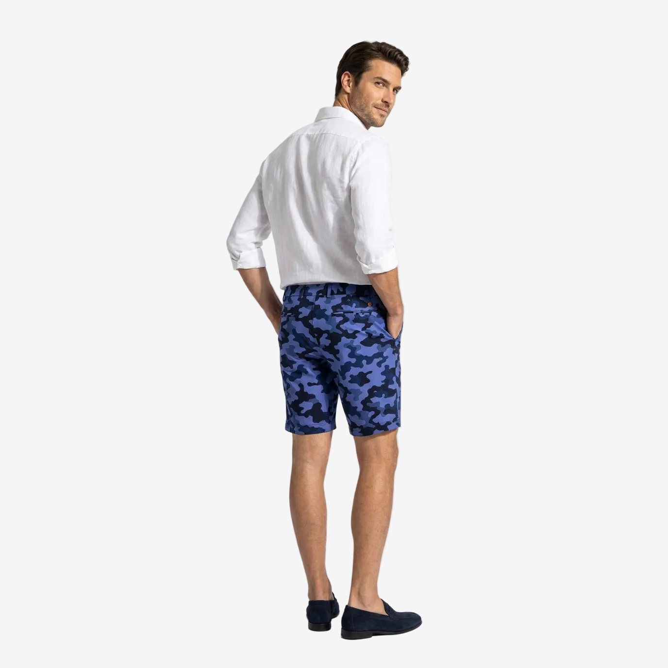 A man wearing blue and navy camouflage shorts, a white button-down shirt, and dark loafers, photographed from the back against a light gray studio background.