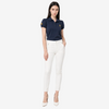 A woman modeling a navy blue short-sleeve polo shirt with a small embroidered logo on the chest and sleeve, paired with white skinny pants and white heels, against a light gray studio background.