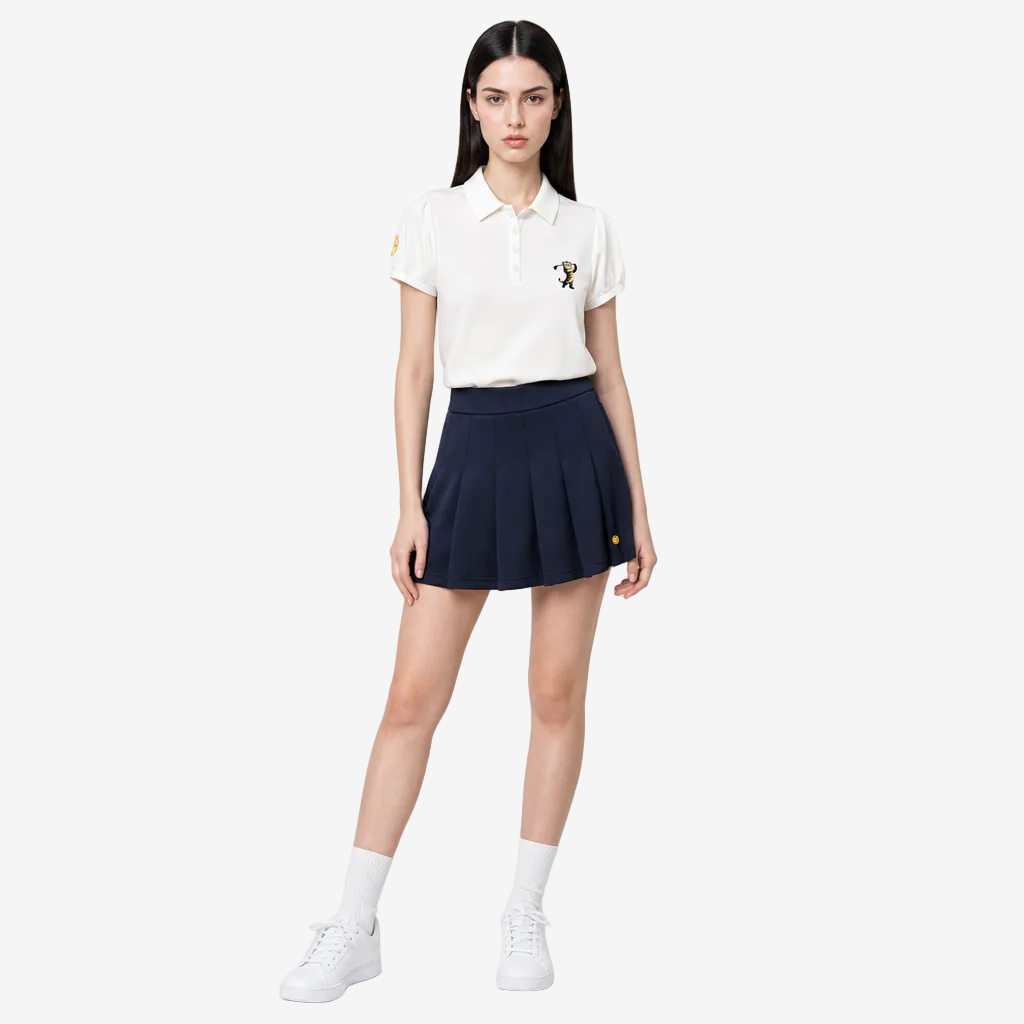 A model stands against a light gray studio background wearing a white short-sleeve polo shirt with small embroidered logos on the chest and sleeve, a dark navy pleated mini skirt, white socks, and white sneakers.