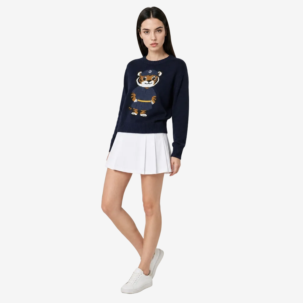 Woman wearing a navy sweater with a tiger design, white pleated skirt, and white sneakers on a light gray background