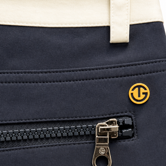 Close-up of a navy blue garment, likely a jacket or coat, featuring a black zipper with a gold-tone pull tab and a small circular gold logo. A cream-colored waistband is visible at the top.