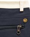 Close-up of a navy blue garment, likely a jacket or coat, featuring a black zipper with a gold-tone pull tab and a small circular gold logo. A cream-colored waistband is visible at the top.