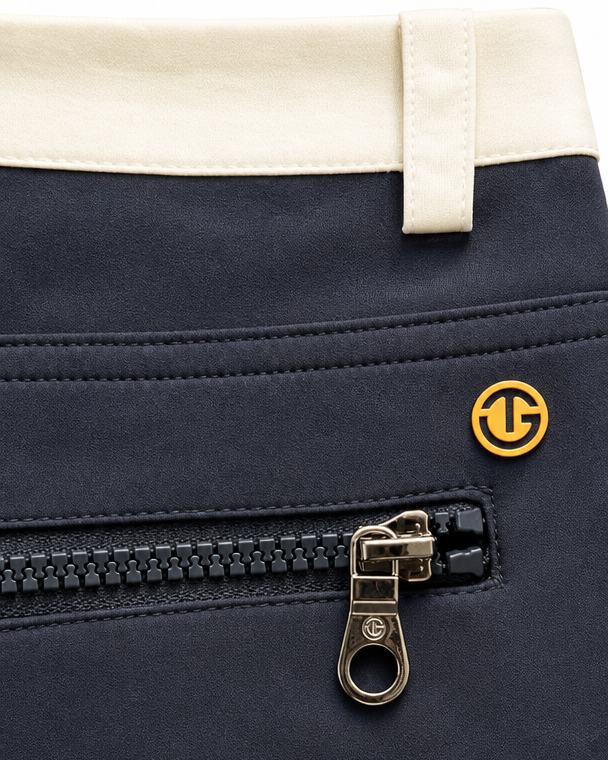 Close-up of a navy blue garment, likely a jacket or coat, featuring a black zipper with a gold-tone pull tab and a small circular gold logo. A cream-colored waistband is visible at the top.