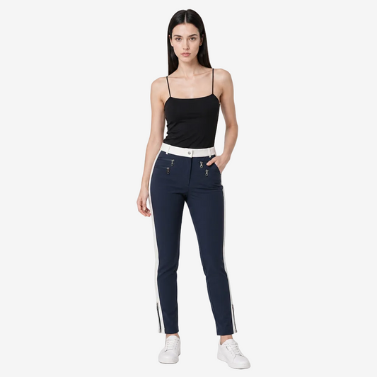 A woman modeling a pair of dark navy blue skinny jeans with white side stripes and a white belt, paired with a black camisole top and white sneakers, against a light gray studio background.