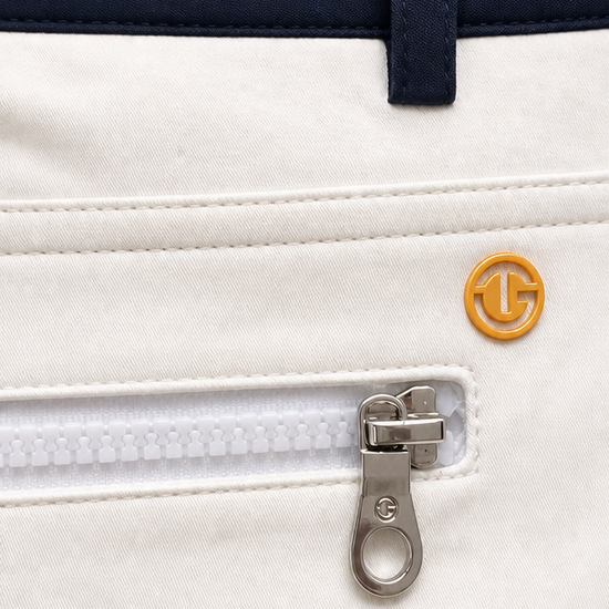 Close-up of a white zippered pocket on a golf jacket, featuring a silver zipper pull with a G-100 logo and a small gold circular G-100 emblem on the front.