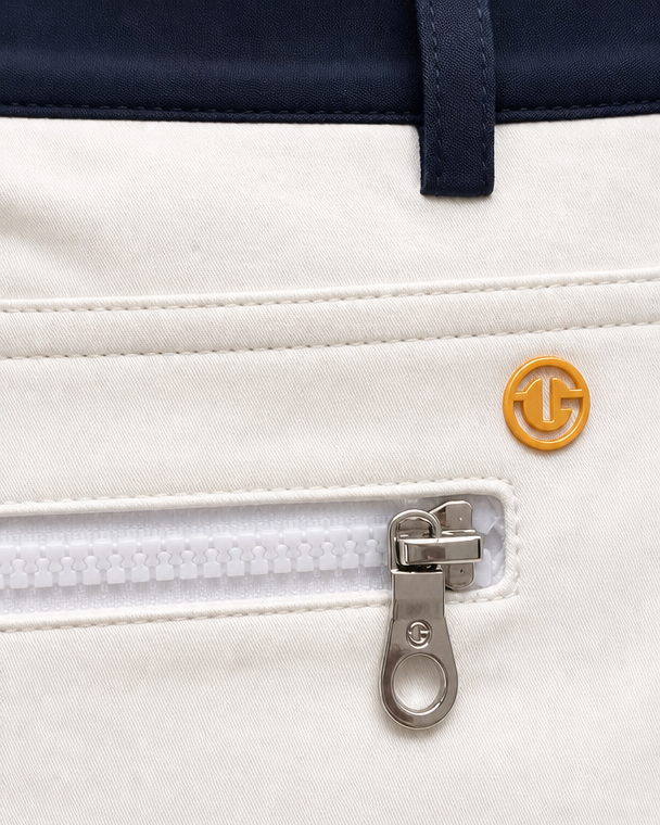 Close-up of a white zippered pocket on a golf jacket, featuring a silver zipper pull with a G-100 logo and a small gold circular G-100 emblem on the front.