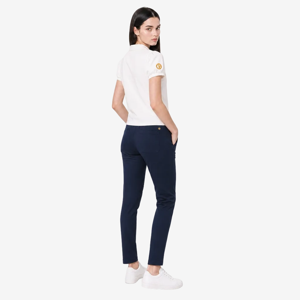 A woman is shown from the back wearing a white short-sleeve polo shirt with a gold circular patch on the sleeve, navy blue slim-fit trousers, and white sneakers, set against a plain light gray studio background.