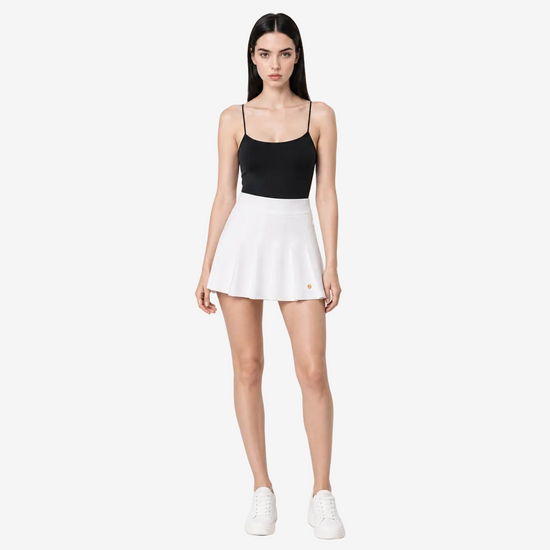 A woman modeling a two-piece athletic set consisting of a black spaghetti-strap tank top and a white tennis skort, paired with white sneakers, against a light gray studio background.