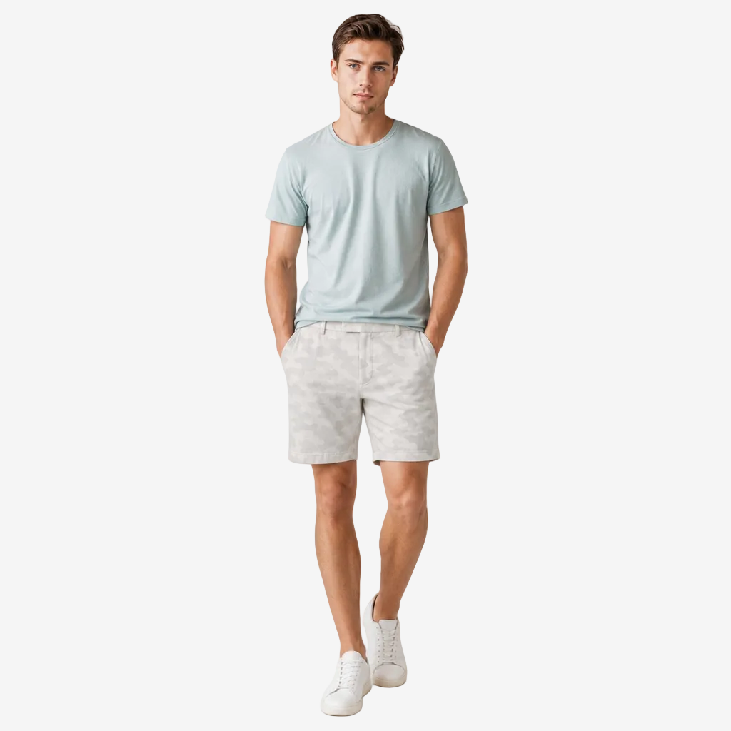 A man modeling a light blue crew-neck t-shirt, light gray patterned shorts, and white sneakers against a plain light gray studio background.