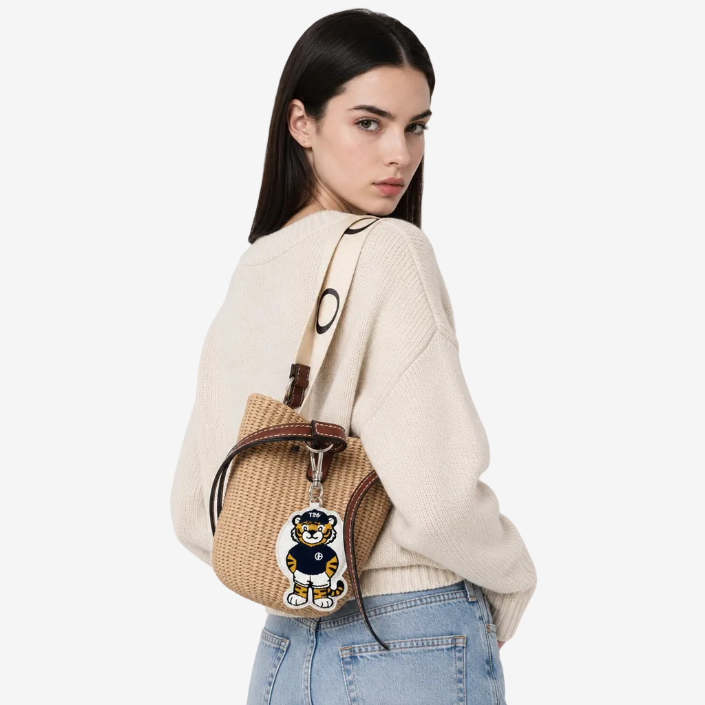 A woman wearing a cream knit sweater and light blue jeans carries a beige woven bucket bag with brown leather trim and a tiger-shaped charm on a plain light gray studio background.