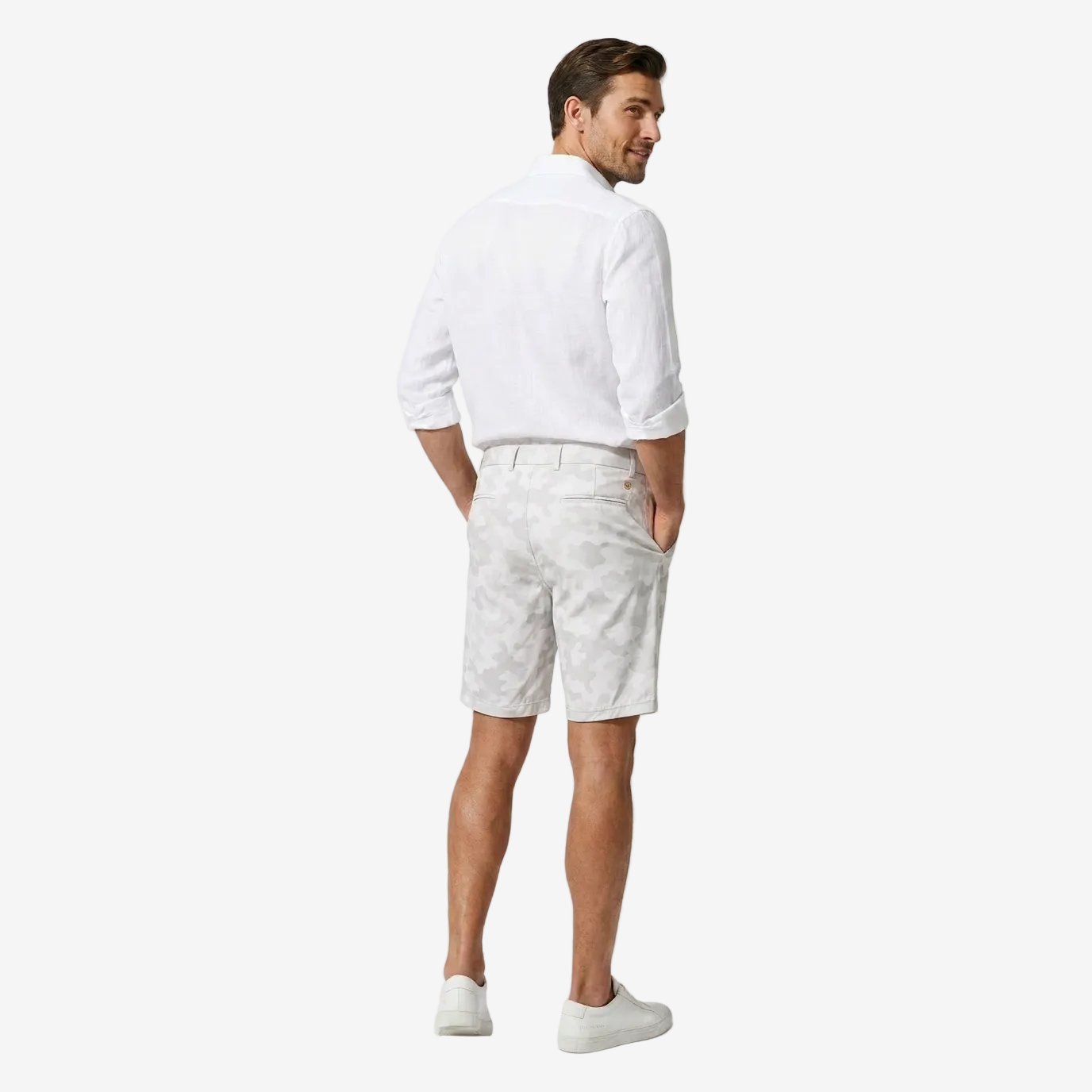 A man wearing a white long-sleeve button-up shirt and light gray patterned shorts, shown from a back-three-quarter view against a light gray studio background.