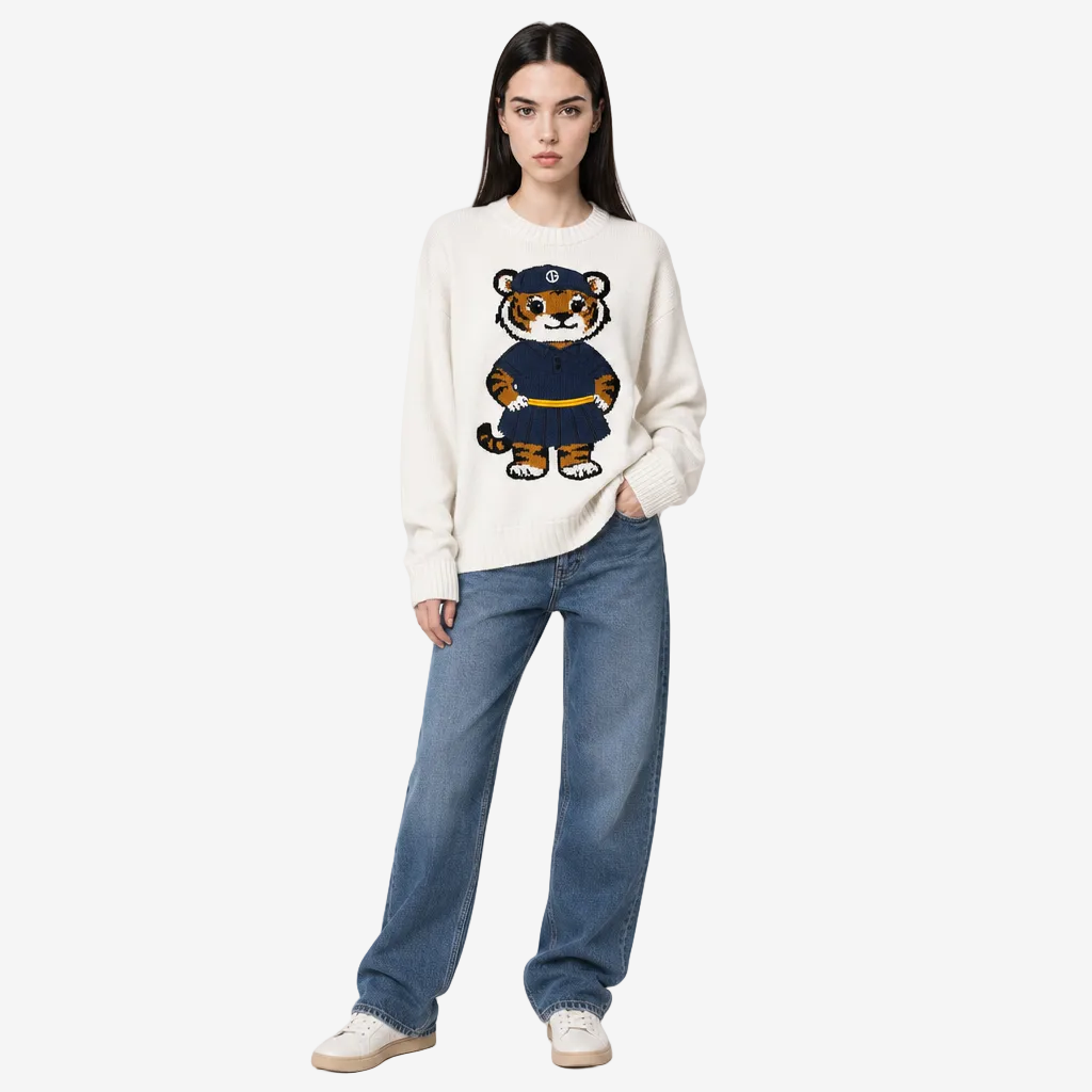 A model stands against a light gray studio background wearing a cream-colored crewneck sweater with a large cartoon tiger graphic on the front, paired with blue jeans and white sneakers.