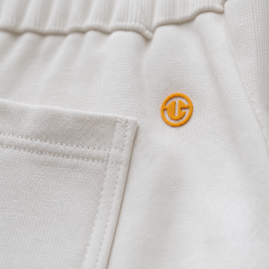 Close-up of off-white knit fabric with a small orange circular logo patch on the side, showing stitched pocket detail and elastic waistband.