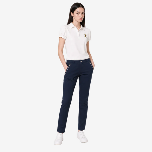 A woman modeling a white short-sleeve polo shirt with a small embroidered logo on the chest, paired with navy blue pants featuring silver zipper pocket details and white sneakers, against a light gray studio background.