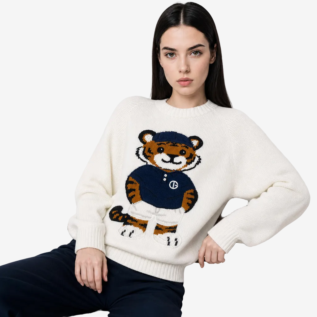 A woman seated on a chair wearing a cream knit sweater with a large embroidered tiger graphic in navy, orange, white, and black. The sweater has a crew neck and ribbed cuffs and hem.