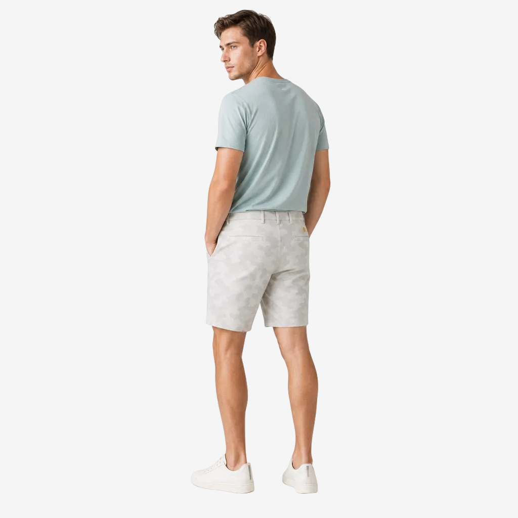 A man wearing a light blue t-shirt, light gray patterned shorts, and white sneakers, photographed from the back against a plain light gray studio background.