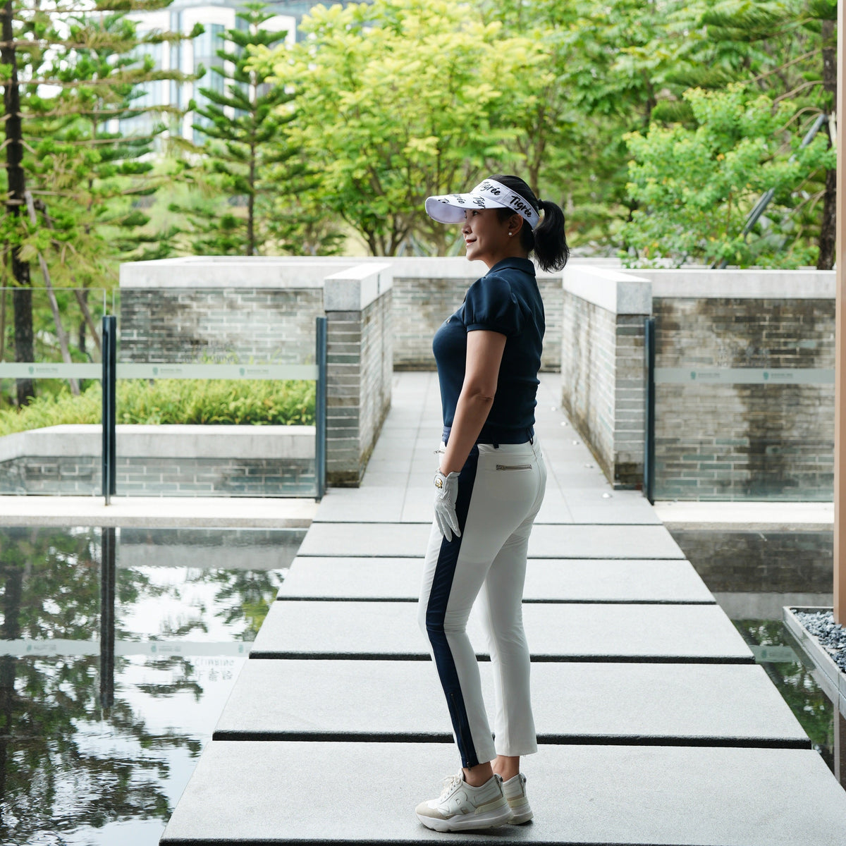 A woman in athletic wear, including a navy blue t-shirt, white and navy striped leggings, and a visor, standing on a modern concrete walkway surrounded by green trees and water.