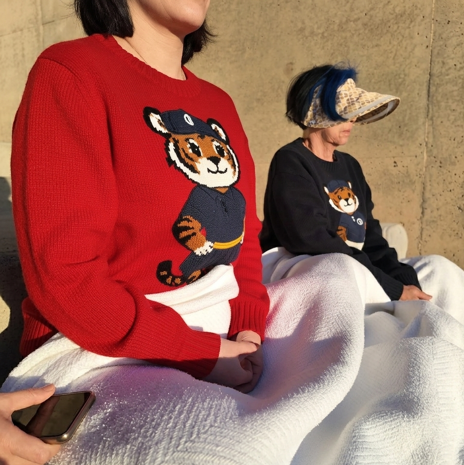 Two people wearing sweaters with a tiger design, sitting on a blanket against a concrete wall.