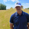 Man wearing a blue cap and shirt standing in a grassy field with a blue sky and white clouds.