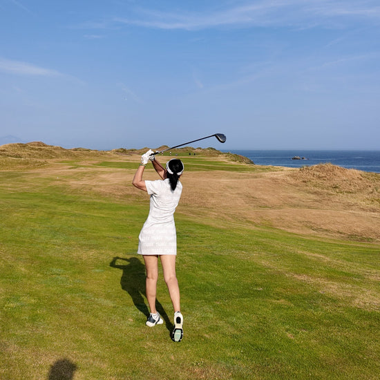 A woman in a white dress swings a golf club on a grassy course under a clear blue sky with the ocean in the distance.
