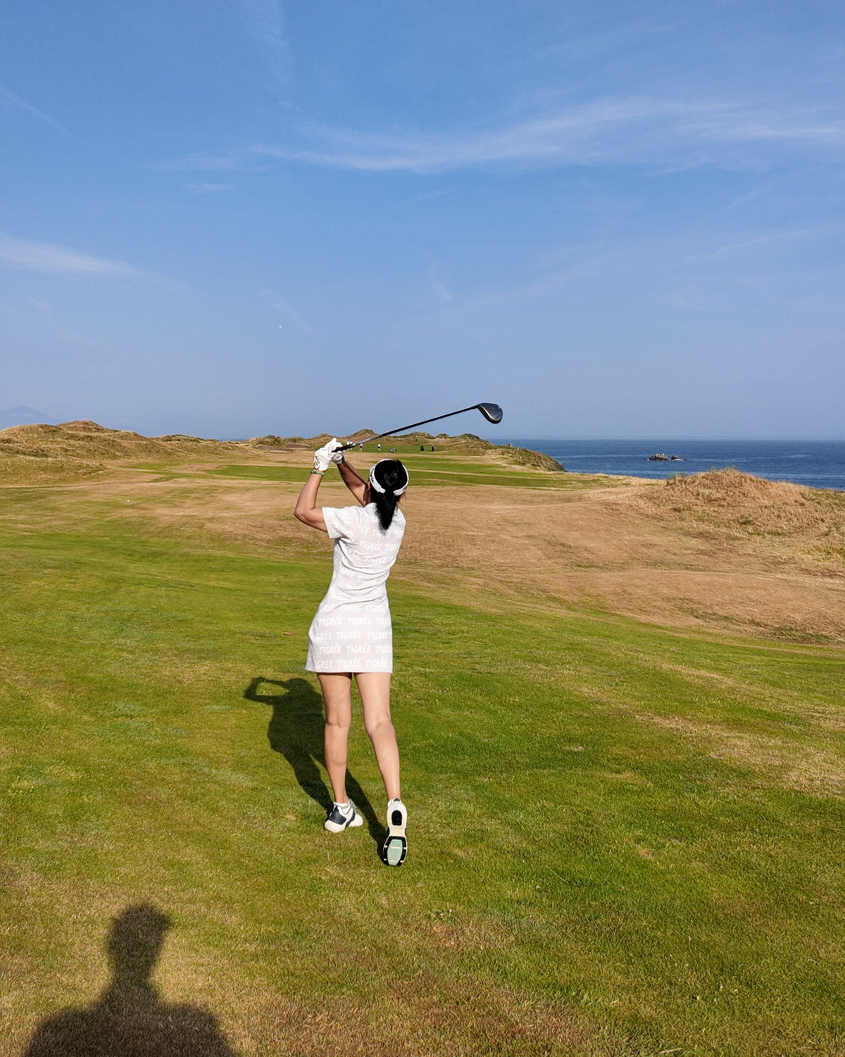 A woman in a white dress swings a golf club on a grassy course under a clear blue sky with the ocean in the distance.