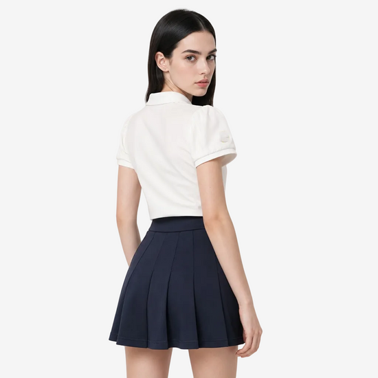 A model is shown from the back wearing a white short-sleeve polo shirt with a small yellow circular logo on the left sleeve, paired with a dark navy pleated mini skirt, against a light gray studio background.