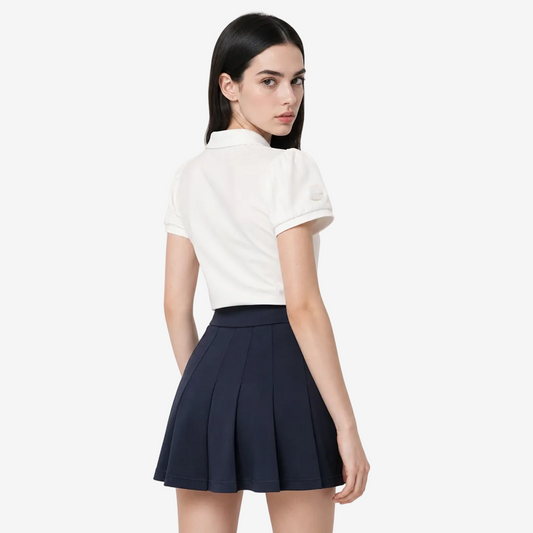 A model is shown from the back wearing a white short-sleeve polo shirt with a small yellow circular logo on the left sleeve, paired with a dark navy pleated mini skirt, against a light gray studio background.