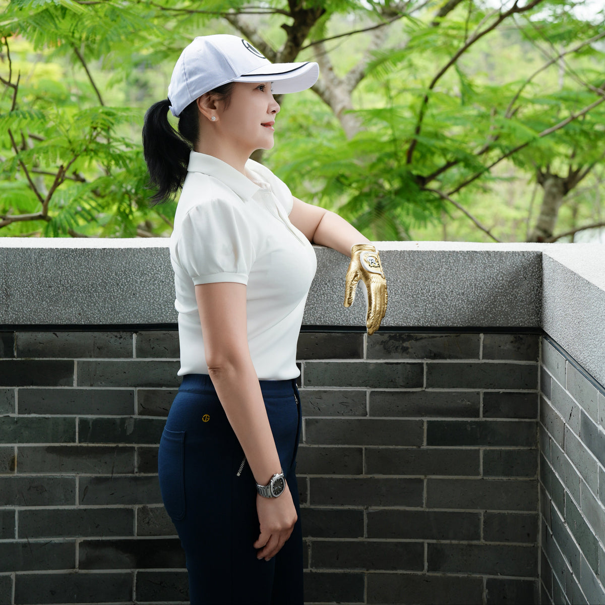A woman in a white polo shirt, navy pants, and a white visor stands outdoors near a gray brick wall, holding a gold golf glove, with green trees in the background.