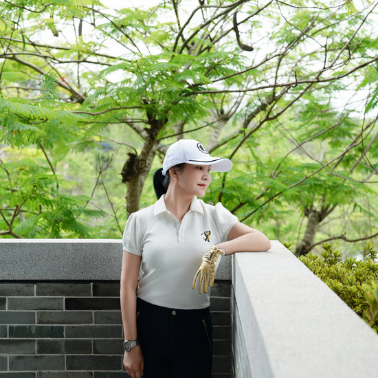 A woman wearing a white polo shirt, black pants, and a white cap stands outdoors leaning on a stone wall, with lush green trees in the background.