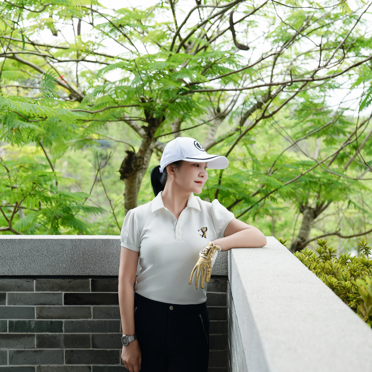 A woman wearing a white polo shirt, black pants, and a white cap stands outdoors leaning on a stone wall, with lush green trees in the background.