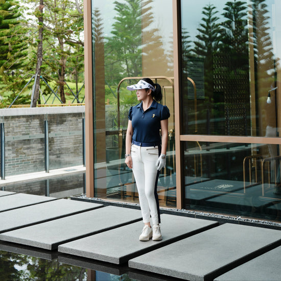 A woman wearing a navy blue polo shirt, white pants, and a white visor stands on a modern outdoor walkway in front of a glass-walled building with trees reflected in the windows.