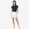 A woman modeling a navy blue polo shirt with a small gold embroidered logo on the chest, paired with a white pleated tennis skirt and white sneakers, against a light gray studio background.